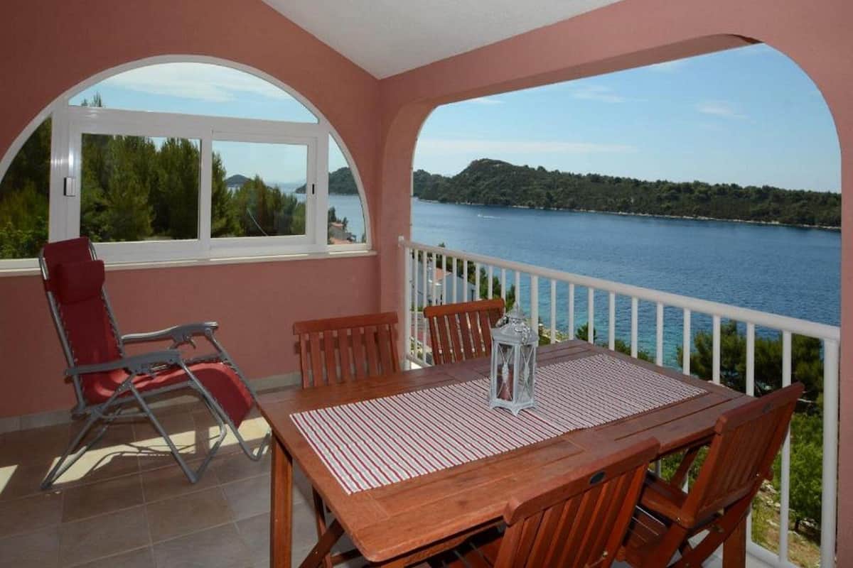 A balcony with a wooden table and chairs, a red chair, and a view of a lake and hills.