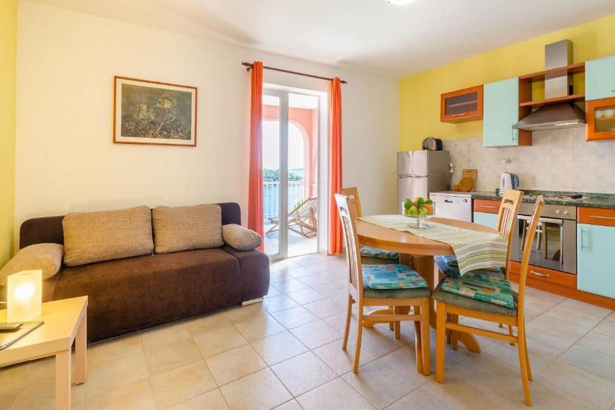 A living room with a sofa, a coffee table, and a dining area with chairs and a table. There is a kitchen area with cabinets and appliances.
