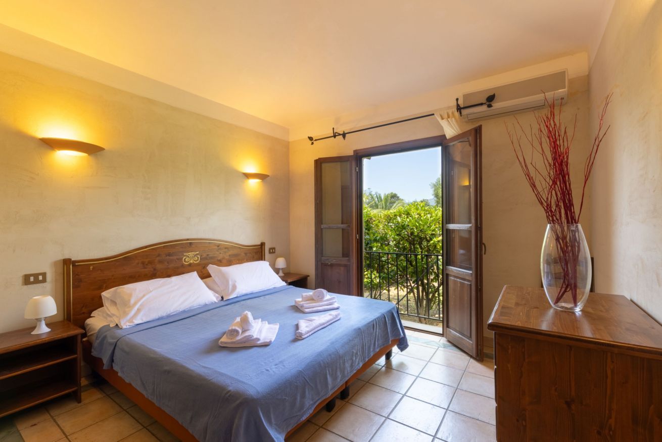 Une chambre à coucher avec un lit, des tables de chevet, une commode, un vase et une vue sur la verdure par une porte ouverte.
