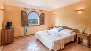 Un dormitorio con cama, televisor, aparador de madera, mesita de noche y ventana con vistas al verdor.