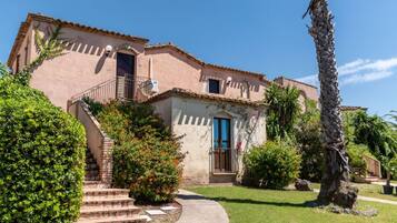 A two-story building with a balcony, a staircase leading to the entrance, and a well-maintained garden with a palm tree and shrubs.