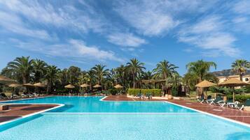 A large, clear swimming pool with lounge chairs and umbrellas, surrounded by palm trees and a clear sky.