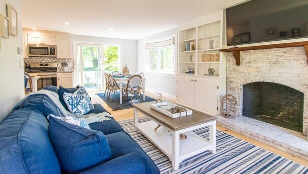 Ein modernes Wohnzimmer mit einem blauen Sofa, einem Couchtisch und einem Kamin. Im Hintergrund ist eine Küche mit weißen Schränken und einer Mikrowelle zu sehen.