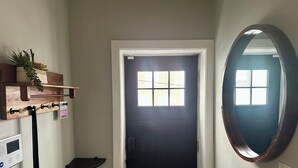 Un pasillo con una puerta negra, un estante de madera con plantas, un termostato en la pared y un espejo redondo.