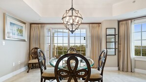 A dining area with a glass table surrounded by ornate chairs, a chandelier, and a view through large windows. - Luxury Condo with Amazing Views Near Attractions (Kissimmee)