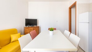 A room with a white table, a yellow sofa, a television on a wooden cabinet, and a white refrigerator.