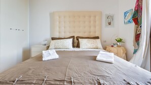 Una cama bien hecha con un cabecero color beige, almohadas blancas y una colcha marrón. Se observa una mesita de noche con una lámpara y un florero con flores.