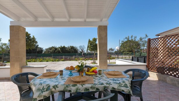 An outdoor patio with a table set for a meal, surrounded by chairs, under a pergola.