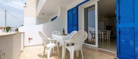 Un balcón exterior con sillas de plástico blanco y una mesa, contraventanas azules y piso de baldosas.