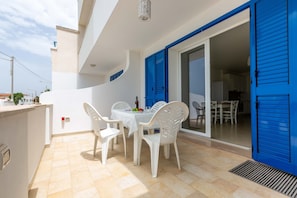 An outdoor balcony with white plastic chairs and a table, blue shutters, and a tiled floor.