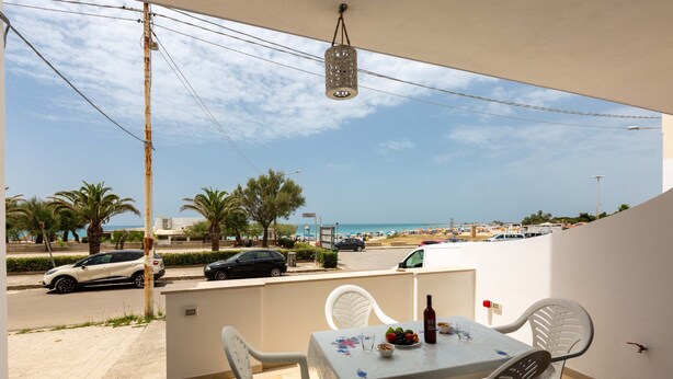 A balcony with a table and chairs, overlooking a beach with palm trees and parked cars.