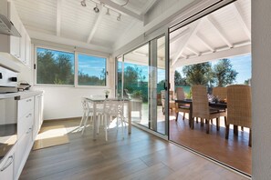 Et moderne køkken med hvide skabe og et spiseområde med fletstole samt en glasdør til en udendørs terrasse.