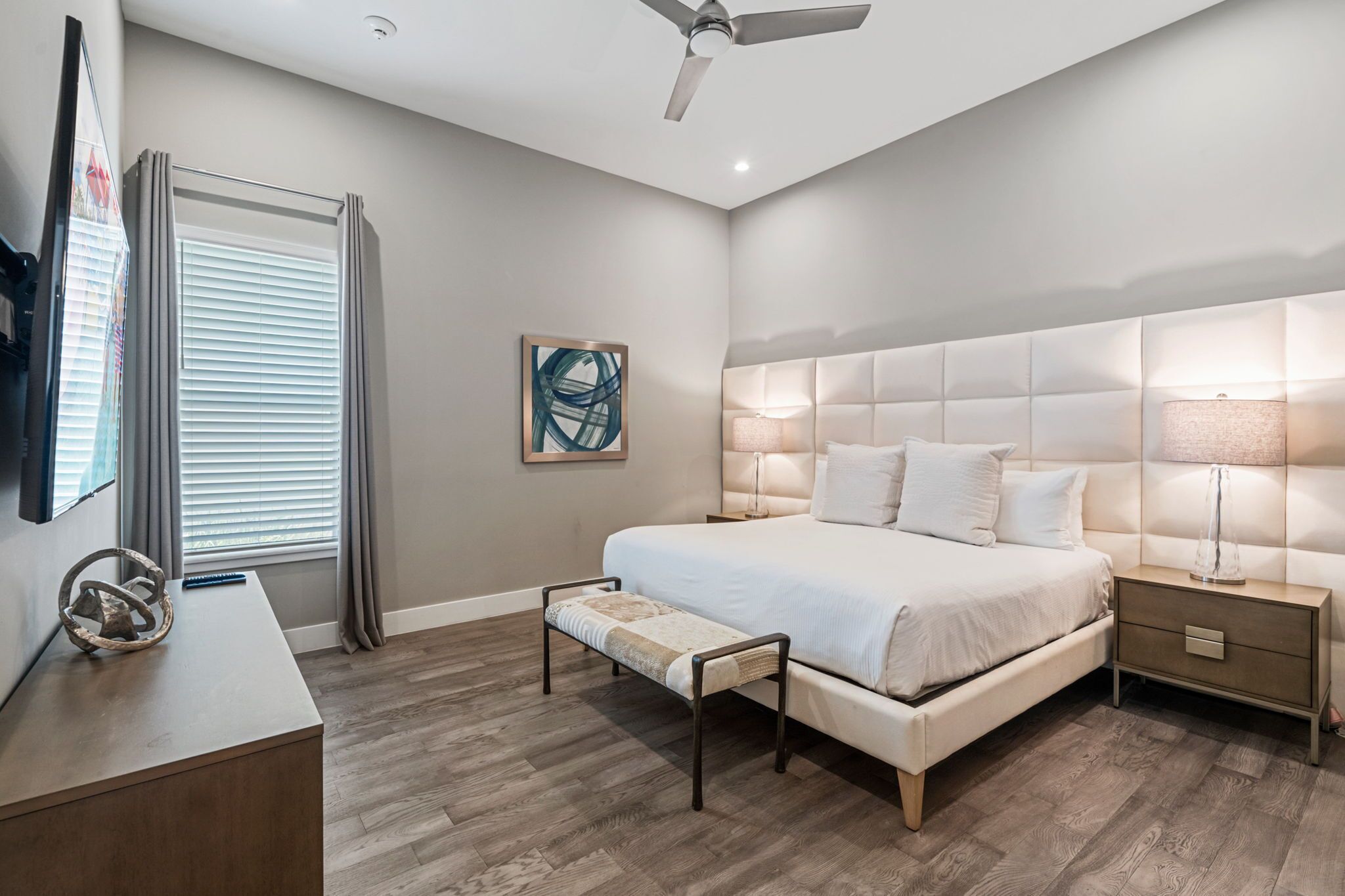 Un dormitorio moderno con una cama grande, lámparas de noche, un banco, un televisor y un ventilador de techo.