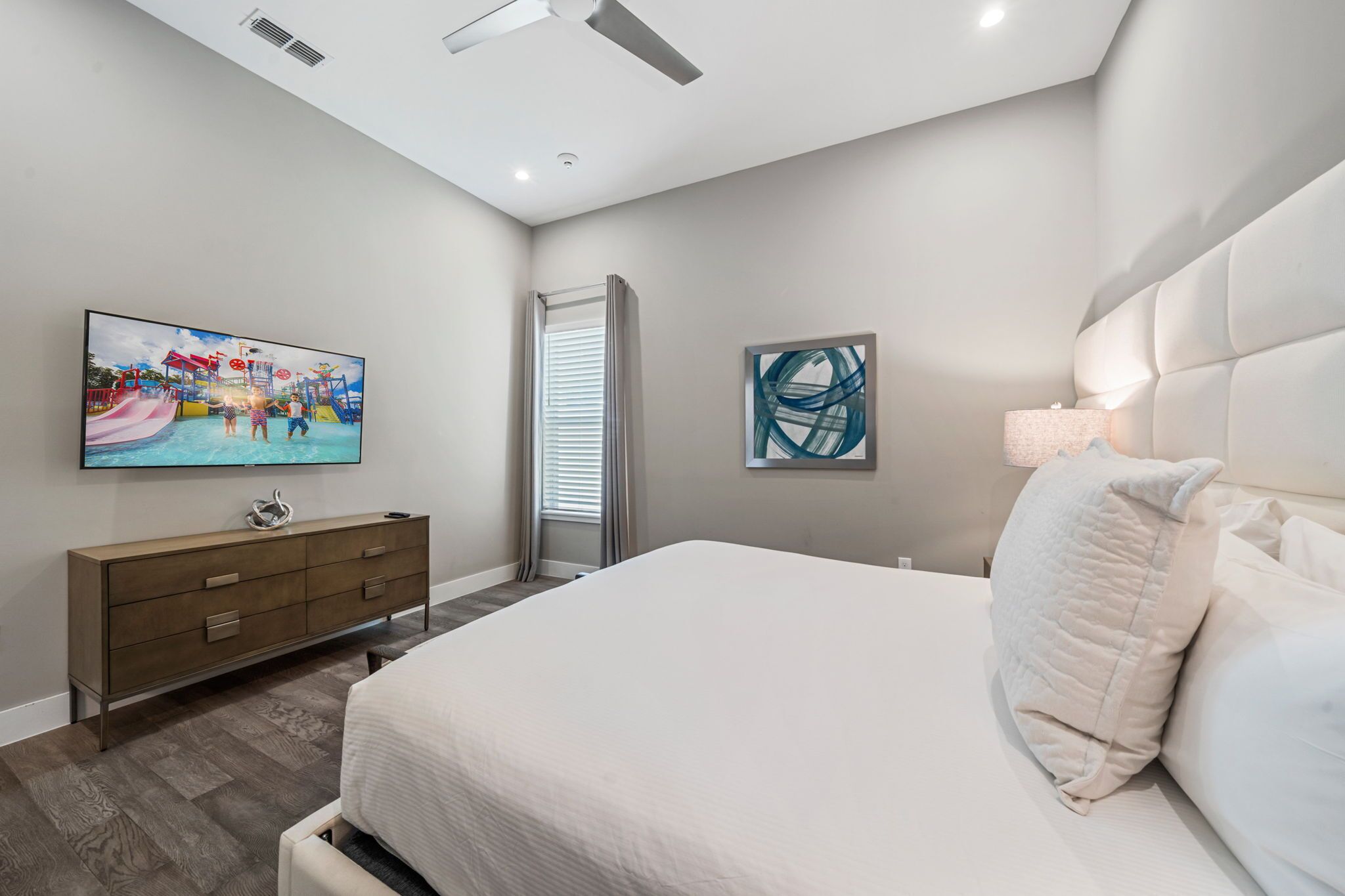 Un dormitorio con cama, televisor montado en la pared, cómoda y ventana con persianas.
