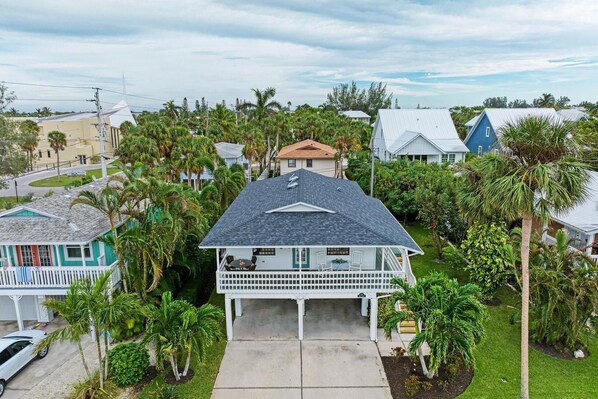 A two-story house with a covered front porch, surrounded by palm trees and other vegetation. - Island Breeze: Steps to Beach, Heated Pool & Spa, Huge Verandah by Anna Maria Life Vacation Rentals (Anna Maria)