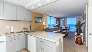 A modern kitchen with white cabinets, a central island, and a dining area with a view of the ocean. - Ilikai Marina #1780, Beautiful Ocean, Sunset and Harbor views! (Honolulu)