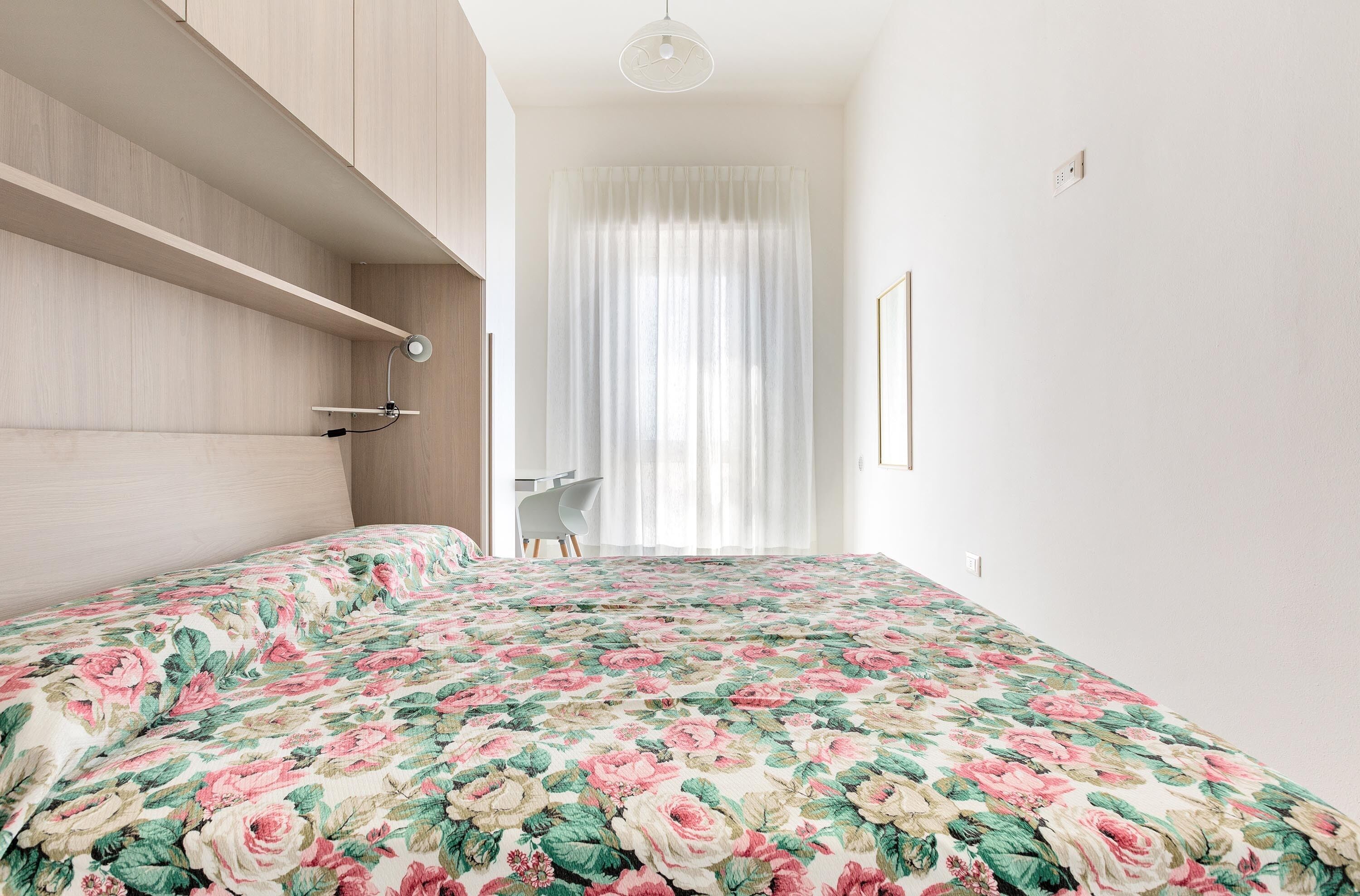 A bedroom with a floral bedspread, a wooden headboard, a bedside table, and a window with curtains.