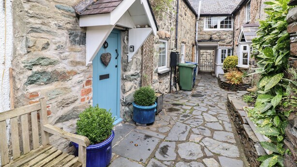 A narrow stone pathway between buildings with a blue door, potted plants, and a stone wall. - 3 Ivy Court (Dolgellau)