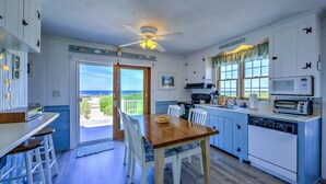 Une cuisine avec une table et des chaises en bois, un ventilateur de plafond et une vue sur l’océan par la porte-fenêtre coulissante.