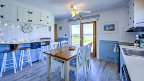 Une cuisine avec une table en bois, des chaises blanches, un ventilateur de plafond et une vue sur l’océan par une porte coulissante.