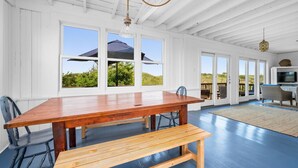Una stanza spaziosa con un grande tavolo di legno e una panca, ampie finestre con vista su un paesaggio verdeggiante e una ventola a soffitto.
