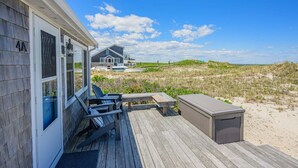Eine Holzterrasse mit Außenmöbeln, einem überdachten Grillbereich und Blick auf eine Strand- und Dünenlandschaft.