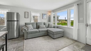 A modern kitchen with a grey sectional sofa, a stainless steel refrigerator, and large windows offering a view of the outside. - Cape Cod Water View Cottage—Steps to Private Beach (East Sandwich)