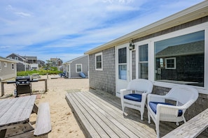 A beachside deck with outdoor furniture, a grill, and a view of neighboring houses.