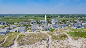 An aerial view of a coastal residential area with houses, sandy terrain, and a lighthouse. - Cape Cod Water View Cottage—Steps to Private Beach (East Sandwich)