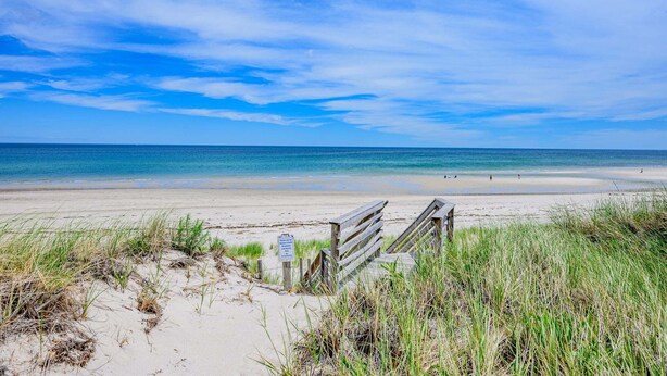 A beach with a wooden boardwalk leading to the water, sandy dunes, and a clear sky. - Cape Cod Water View Cottage—Steps to Private Beach (East Sandwich)