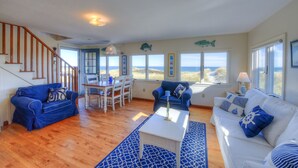 Ein Wohnzimmer mit einer Holztreppe, zwei blauen Sofas, einem weißen Sofa, einem Esstisch mit Stühlen und großen Fenstern mit Blick auf den Strand.