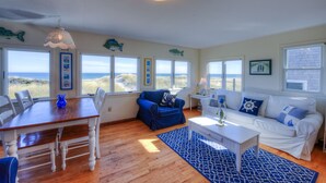 Ein geräumiges Wohnzimmer mit einem Holzesstisch, einem blauen Sofa und einem weißen Couchen. Mehrere Fenster bieten Blick auf den Strand.