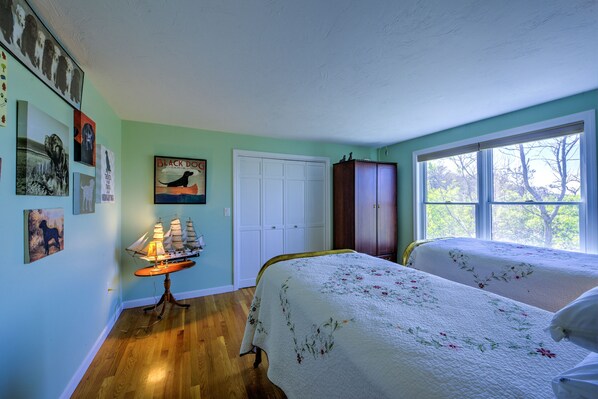 A bedroom with two beds, a wooden floor, a window with a view of trees, a door, and a cabinet.
