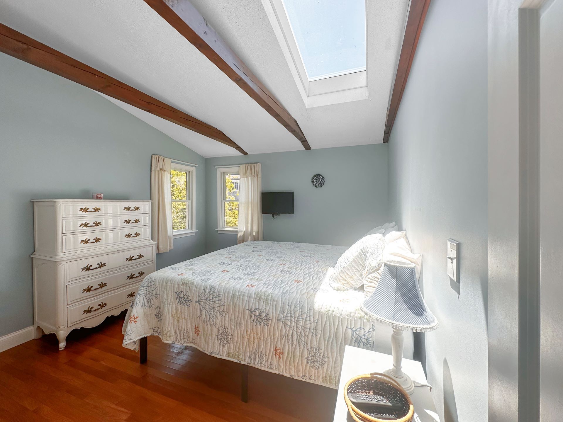 A bedroom with a skylight, a bed with a floral bedspread, a dresser with multiple drawers, two windows with curtains, and a wall-mounted TV.