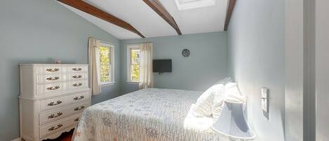 A bedroom with a skylight, a bed with a floral bedspread, a dresser with multiple drawers, two windows with curtains, and a wall-mounted TV.