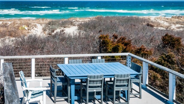 A deck with a table and chairs overlooking a beach with ocean waves. - Beachside Bliss with Skylights & Fireplace (East Sandwich)