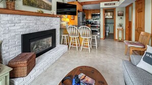 A spacious living area with a fireplace, a dining table with chairs, and a kitchen in the background. - Beachside Bliss with Skylights & Fireplace (East Sandwich)