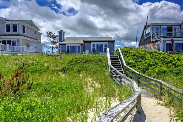 A coastal property with a wooden boardwalk leading to a house with a large porch and a second-story balcony.