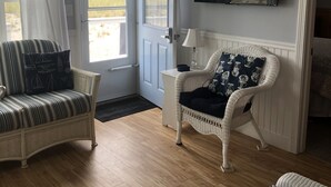 A room with a wooden floor, a white wicker chair with a dark cushion, a striped sofa, and a television mounted on the wall. - Oceanfront Cottage—Private Beach & Bay Views (East Sandwich)