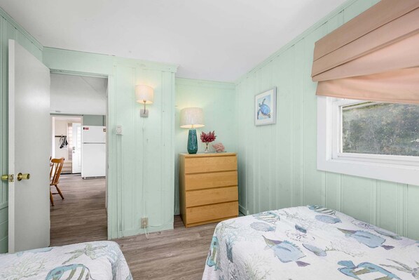 Une chambre avec deux lits, une commode, des appliques murales, une fenêtre avec des stores et une porte donnant sur une autre pièce.