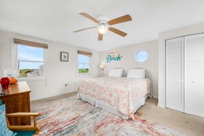 A bedroom with a bed, a ceiling fan, a window with a view, a wall with a 'Life is better at the Beach' sign, and a white closet.