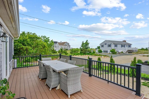 A wooden deck with a table and chairs, surrounded by a black railing, overlooking a residential area with houses and greenery.
