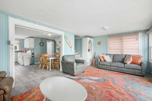 Un salon avec un canapé gris, une table basse blanche et un tapis coloré. On y trouve également un coin repas avec des chaises et une table en bois, ainsi qu'une cuisine avec des meubles blancs et un lave-linge.