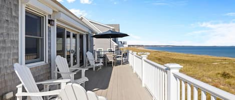 A deck with white Adirondack chairs, a table, and an umbrella, overlooking a beach.