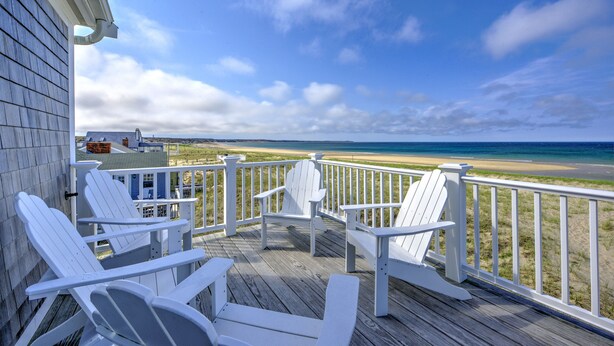 Eine Holzterrasse mit weißen Adirondack-Stühlen mit Blick auf einen Strand.