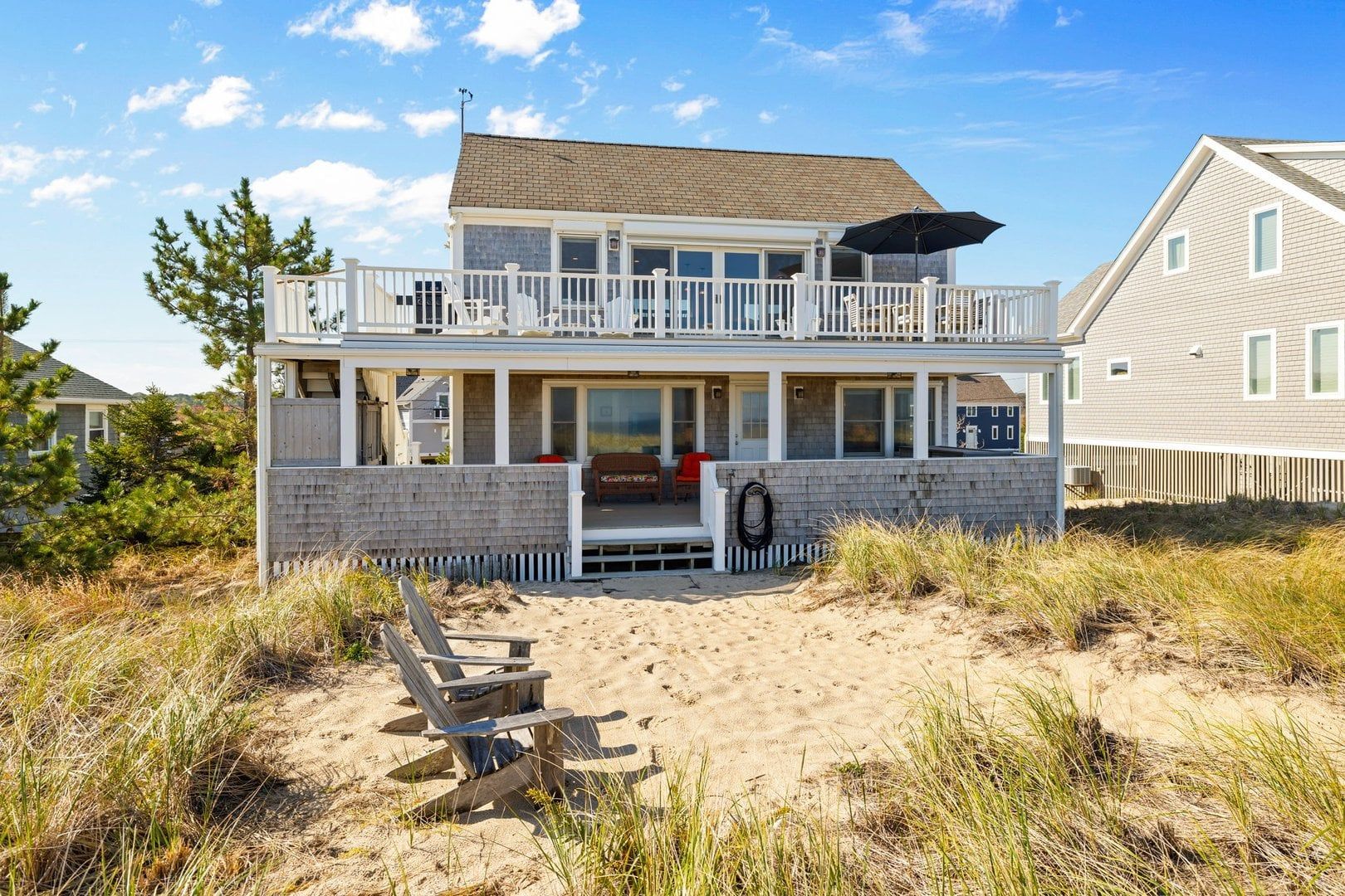 A two-story beach house with a balcony, a covered porch, and a sandy front yard.