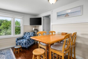 A room with a wooden dining table, four chairs, a television mounted on the wall, and a window with a view of greenery.