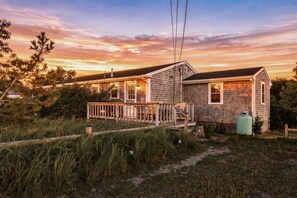 Een houten huis met twee verdiepingen en een terras, omringd door gras en bomen, tijdens de zonsondergang.