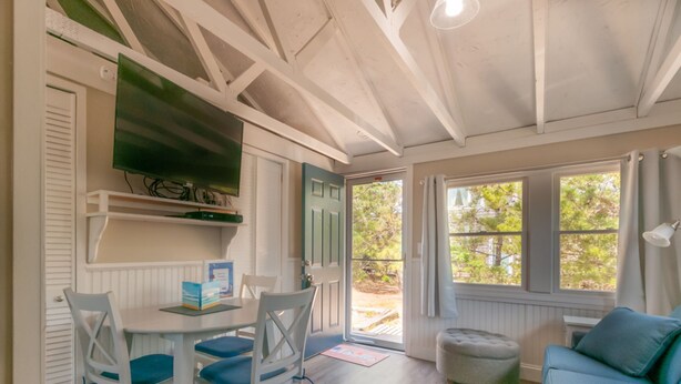 Ein gemütliches Zimmer mit Esstisch und Stühlen, einer Couch, einem an der Wand befestigten Fernseher und einem Fenster mit Blick auf Bäume.