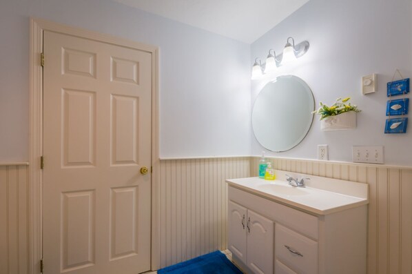 A bathroom with a white door, a round mirror, a white sink with a faucet, and a blue bath mat. - Oceanview Retreat w/ Private Beach Access (Bourne)